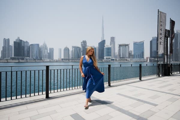 Elegant woman in a blue dress posing by Dubai's iconic skyline on a sunny day.