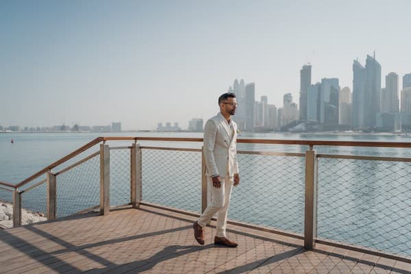 A stylish man in a suit walking along the waterfront with Dubai's skyline in the background.